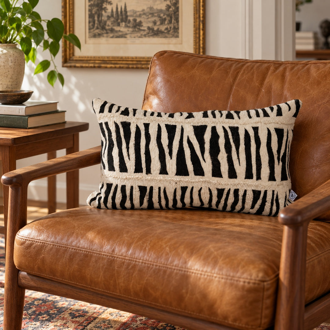 Coussin IDUBE 30 × 50 cm en bogolan zébré noir et écru sur un fauteuil vintage en cuir caramel.