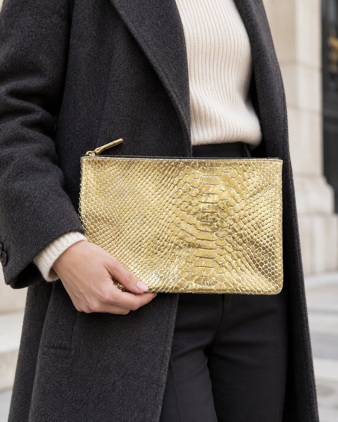 LOVE-Etui von Isabelle Varin aus goldfarbenem Leder mit Alligator-Prägung, in der Hand getragen zu einem anthrazitfarbenen Mantel und einem ecrufarbenen Pullover.