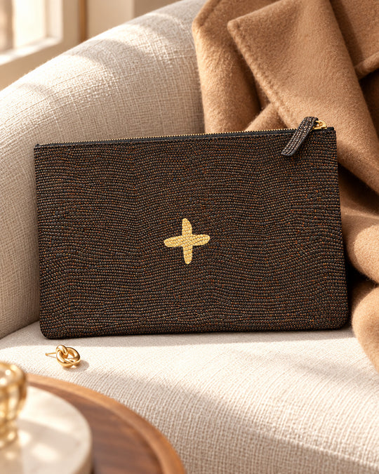 LOVE-Etui von Isabelle Varin aus schimmerndem braunem Leder, mit goldenem Kreuz, auf einem Leinensessel neben einem camelfarbenen Mantel.