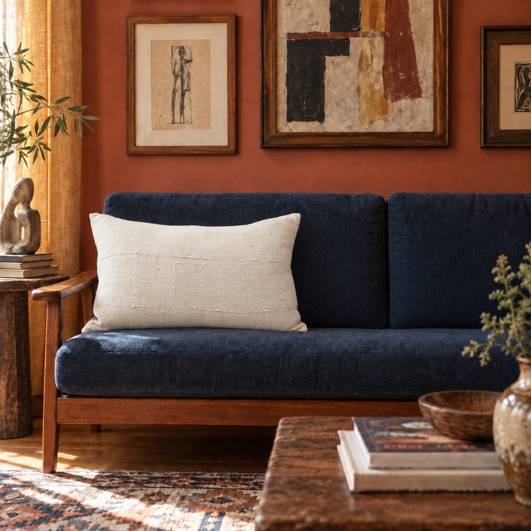Rechthoekig ecru MSINGI kussen op een blauwe bank met houten frame, in een woonkamer met terracottakleurige muren en ingelijste kunstwerken.