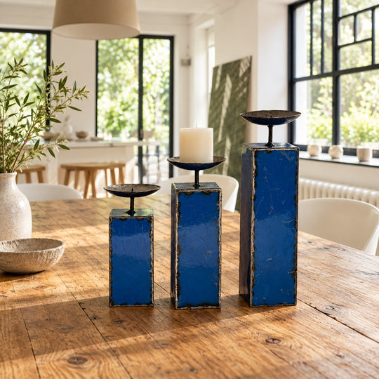 Trois bougeoirs NURU en métal recyclé bleu sur une table en bois dans une salle à manger lumineuse.