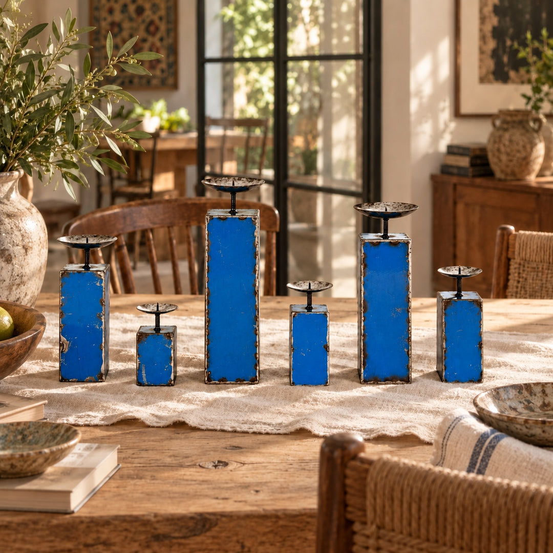 Six bougeoirs NURU bleus en métal recyclé, de tailles variées, sur une table en bois patiné avec un chemin de table en lin écru.