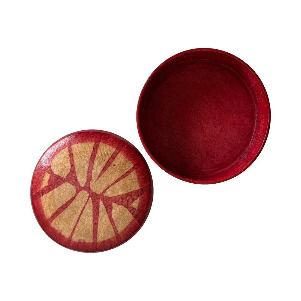 Boîte ronde en cuir rouge, ouverte sur un intérieur uni, avec son couvercle à motif rayonnant beige à côté.