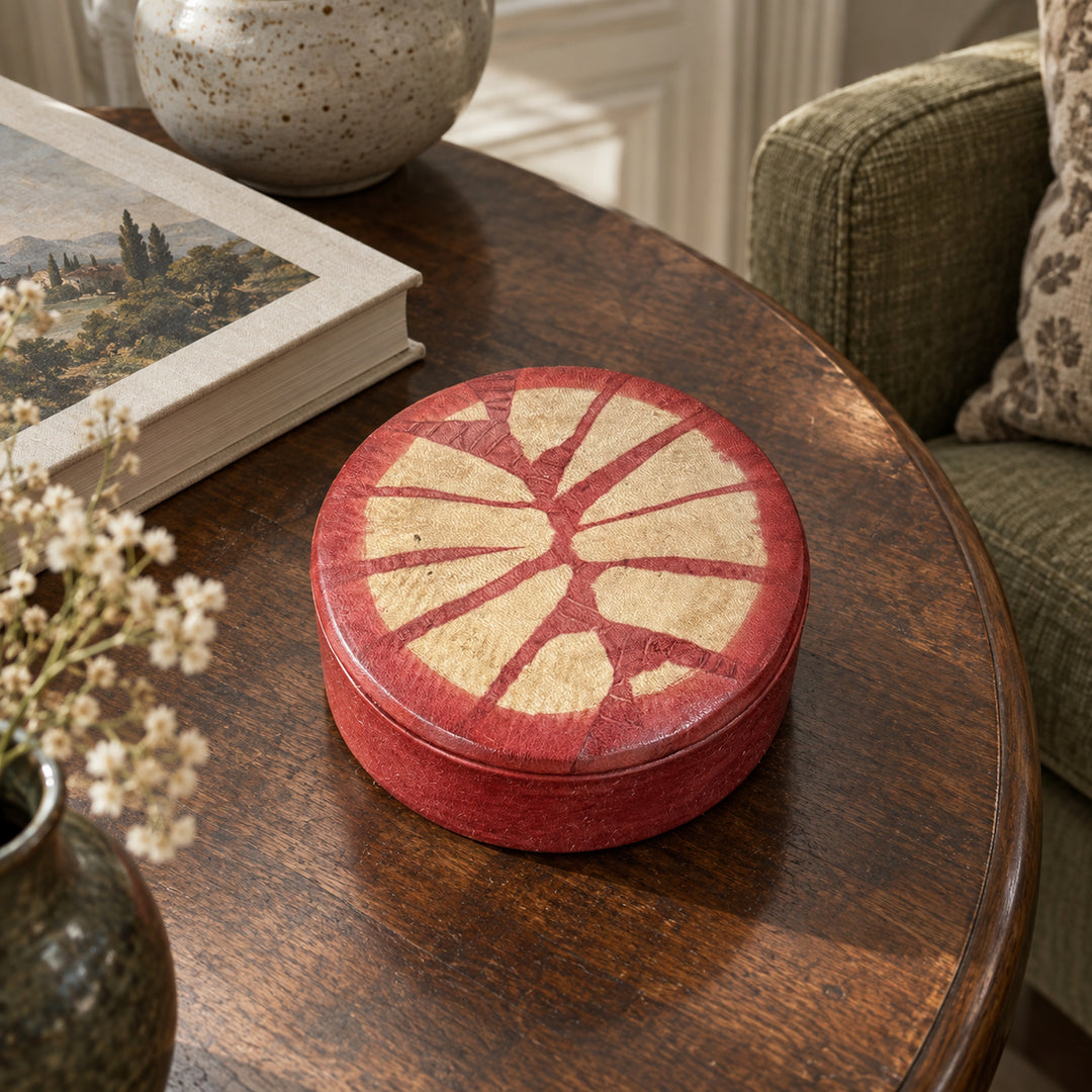 Boîte ronde en cuir rouge à motif rayonnant beige, sur une table ronde en bois près d’un livre et d’un fauteuil vert.
