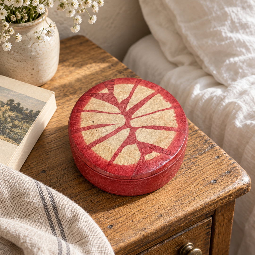 Boîte ronde en cuir rouge à motif rayonnant beige, sur une table de chevet en bois près d’un livre et d’un lit au linge blanc.