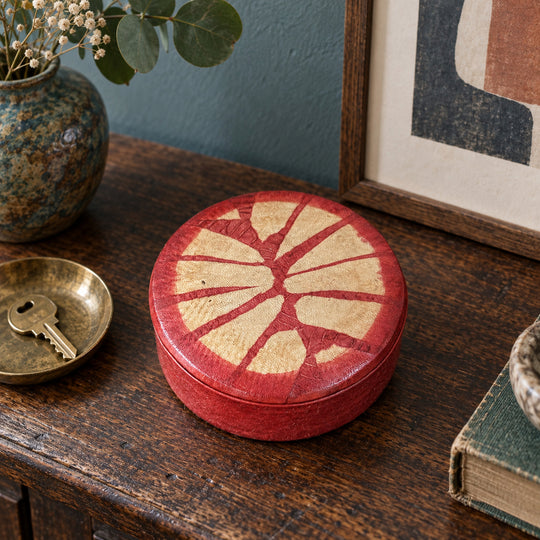 Boîte ronde en cuir rouge à motif rayonnant beige, sur une console en bois devant une œuvre encadrée, près d’une coupelle avec une clé.