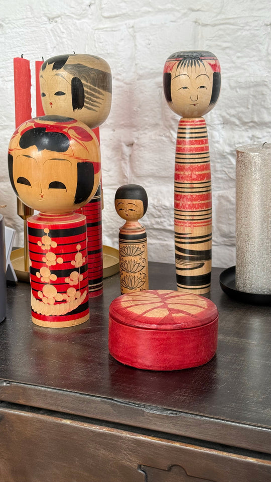 Boîte ronde rouge à motif rayonnant beige, posée sur un meuble en bois devant des poupées japonaises kokeshi.