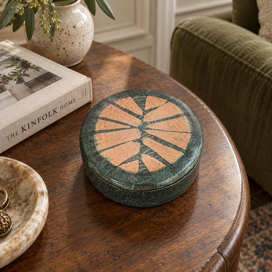 Boîte ronde en cuir vert foncé à motif rayonnant saumon, sur une table ronde en bois près d’un livre et d’un fauteuil vert.