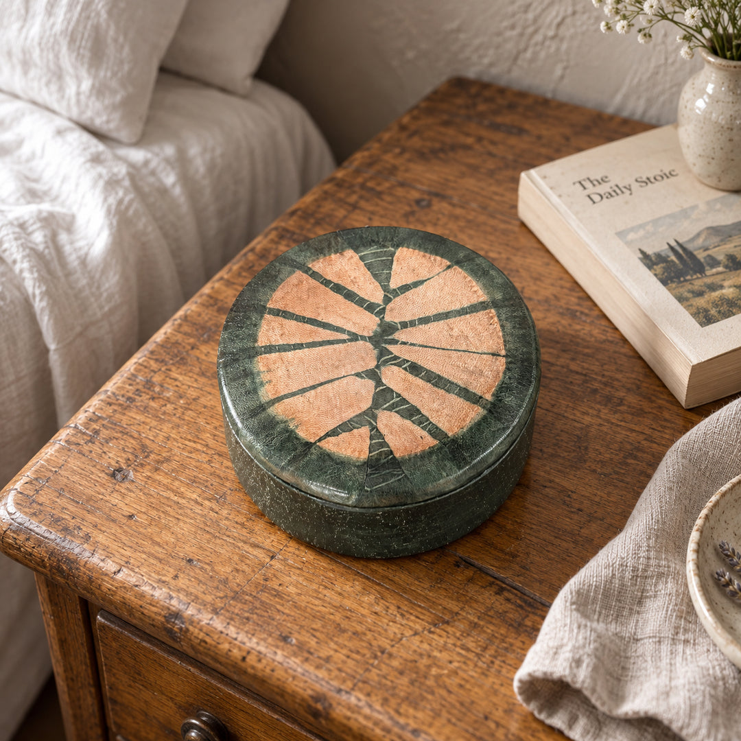 Boîte ronde en cuir vert foncé à motif rayonnant saumon, sur une table de chevet en bois près d’un livre et d’un lit au linge blanc.