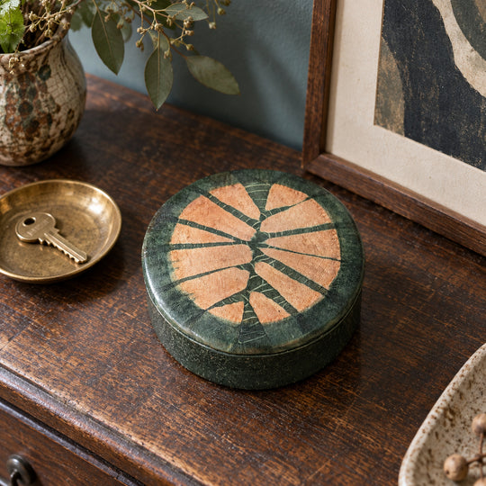 Boîte ronde en cuir vert foncé à motif rayonnant saumon, sur une console en bois devant une œuvre encadrée, près d’une coupelle avec une clé.