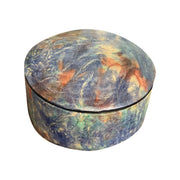 Boîte ronde en cuir marbré bleu, turquoise et orange, fermée et vue de trois quarts sur fond blanc.