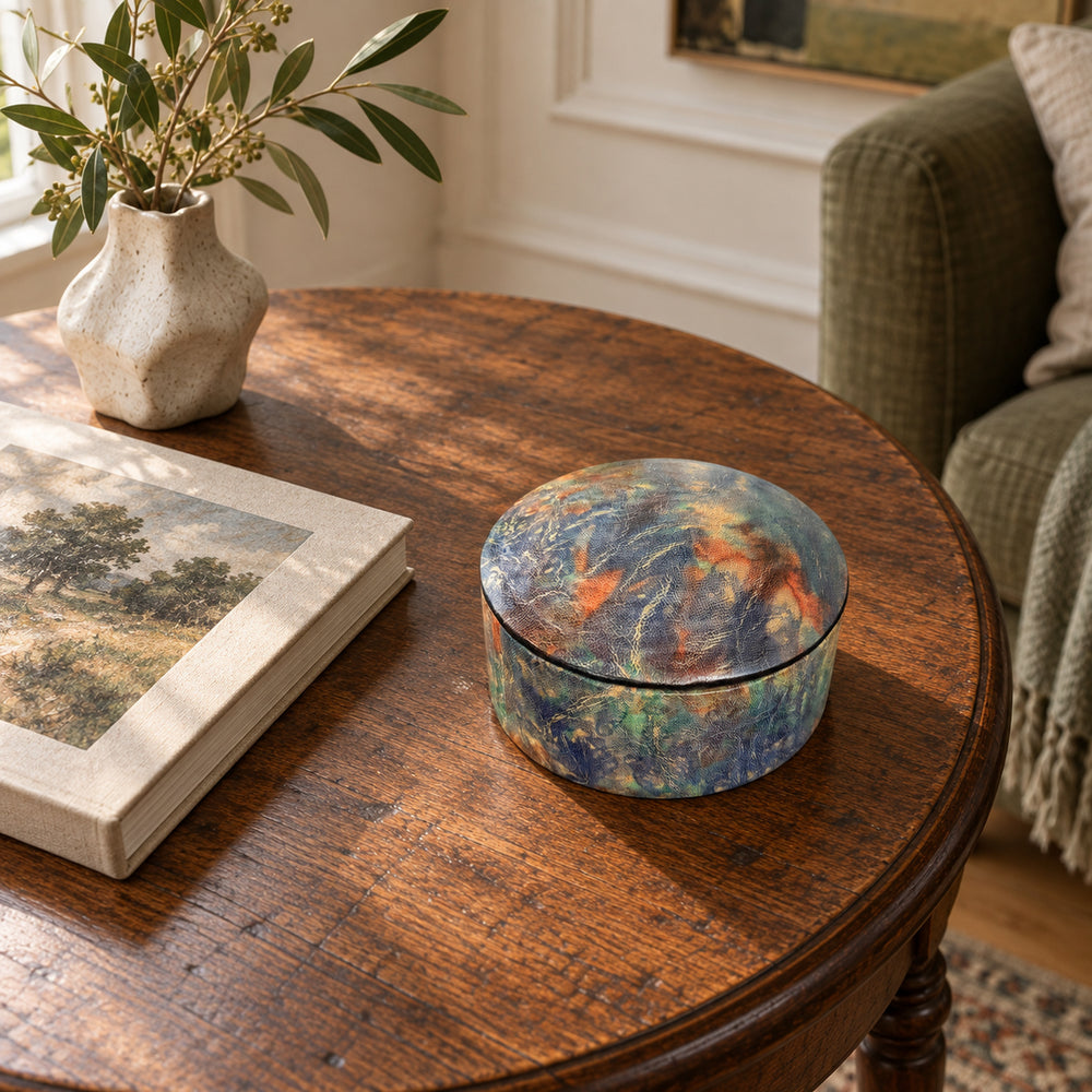 Boîte ronde en cuir marbré bleu, turquoise et orange, sur une table ronde en bois près d'un livre et d'un fauteuil vert.