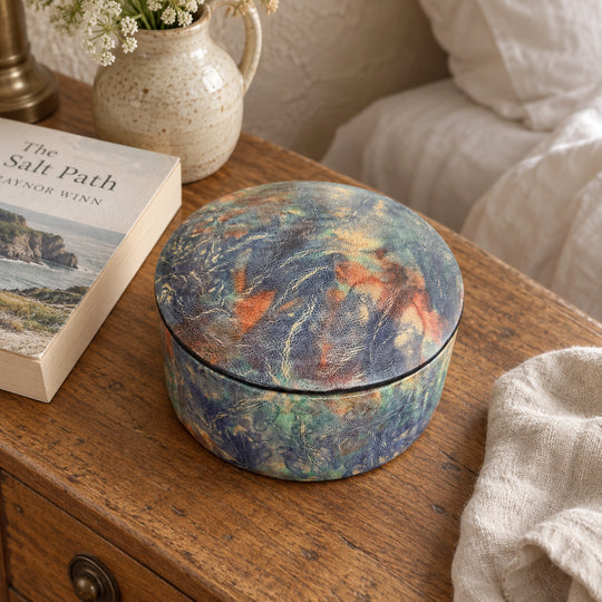 Boîte ronde en cuir marbré bleu, turquoise et orange, sur une table de chevet en bois près d'un livre et d'un lit au linge blanc.