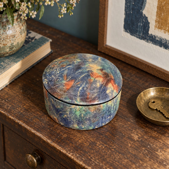 Boîte ronde en cuir marbré bleu, turquoise et orange, sur une console en bois devant une œuvre encadrée, près d'une coupelle avec une clé.