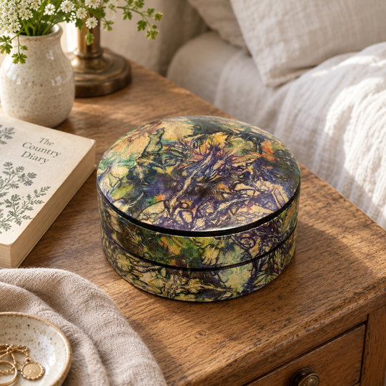 Boîte ronde en cuir marbré violet, vert et ocre, sur une table de chevet en bois près d'un livre et d'un lit au linge blanc.