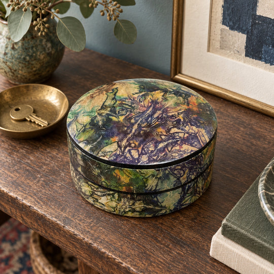 Boîte ronde en cuir marbré violet, vert et ocre, sur une console en bois devant une œuvre encadrée, près d'une coupelle avec une clé.