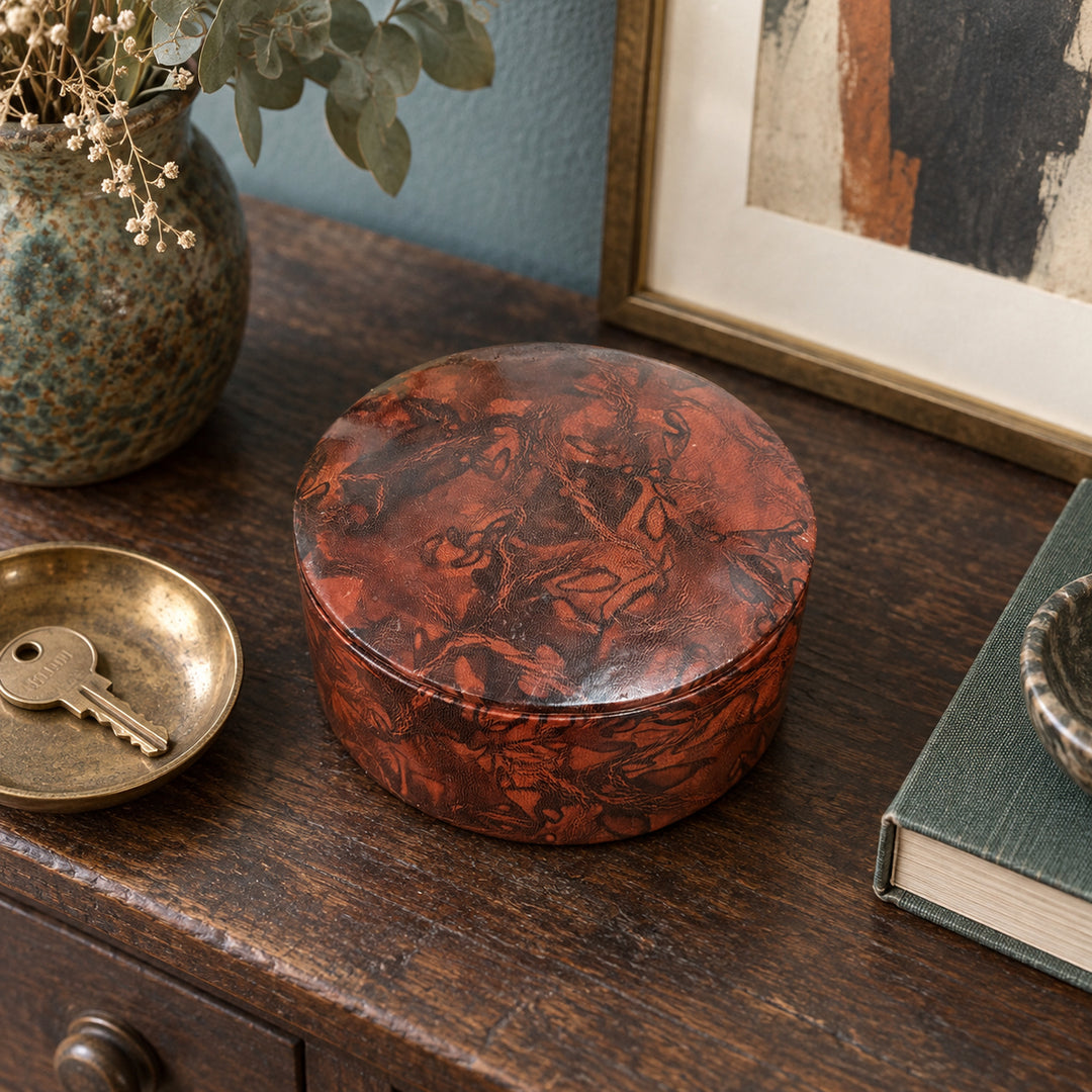 Boîte ronde en cuir marbré rouge rouille et brun, sur une console en bois devant une œuvre encadrée, près d'une coupelle avec une clé.