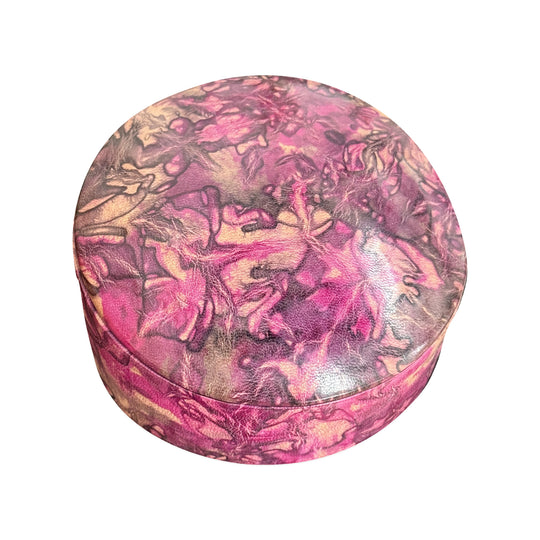 Boîte ronde en cuir marbré rose fuchsia, prune et beige, fermée et vue de trois quarts sur fond blanc.