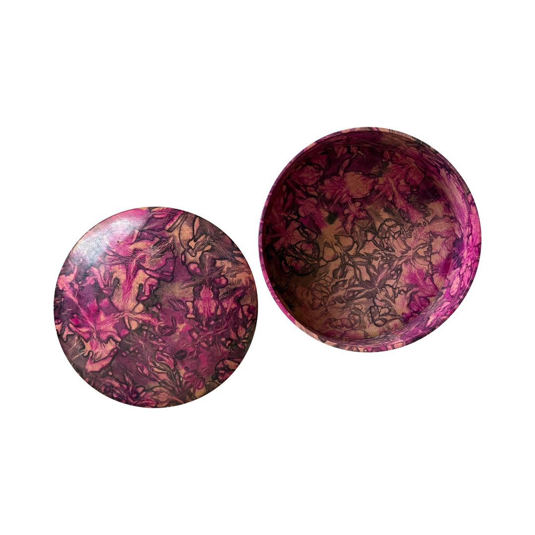 Boîte ronde en cuir marbré rose fuchsia, prune et beige, ouverte avec son couvercle posé à côté sur fond blanc.