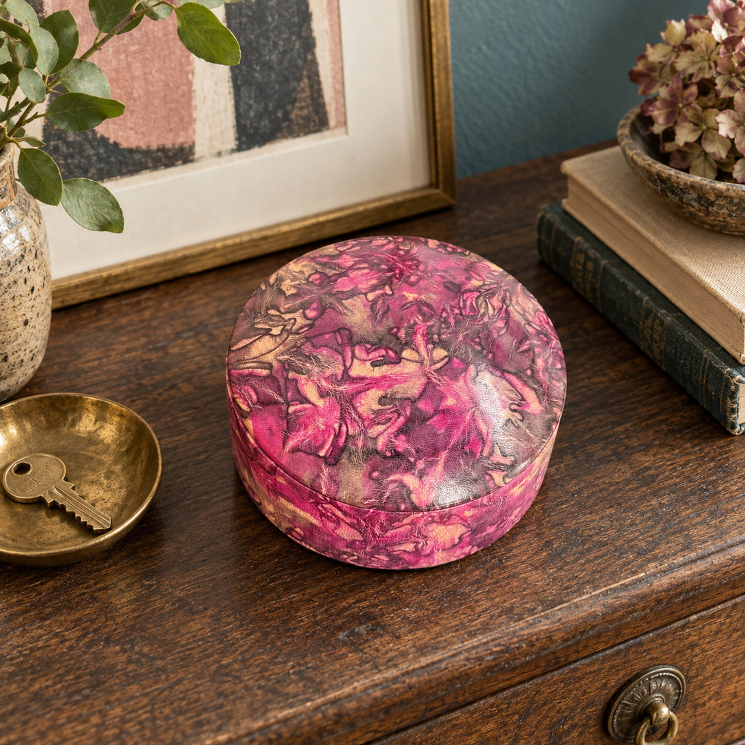 Boîte ronde en cuir marbré rose fuchsia, prune et beige, sur une console en bois devant une œuvre encadrée, près d'une coupelle avec une clé.