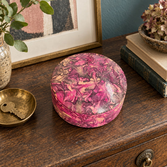 Boîte ronde en cuir marbré rose fuchsia, prune et beige, sur une console en bois devant une œuvre encadrée, près d'une coupelle avec une clé.