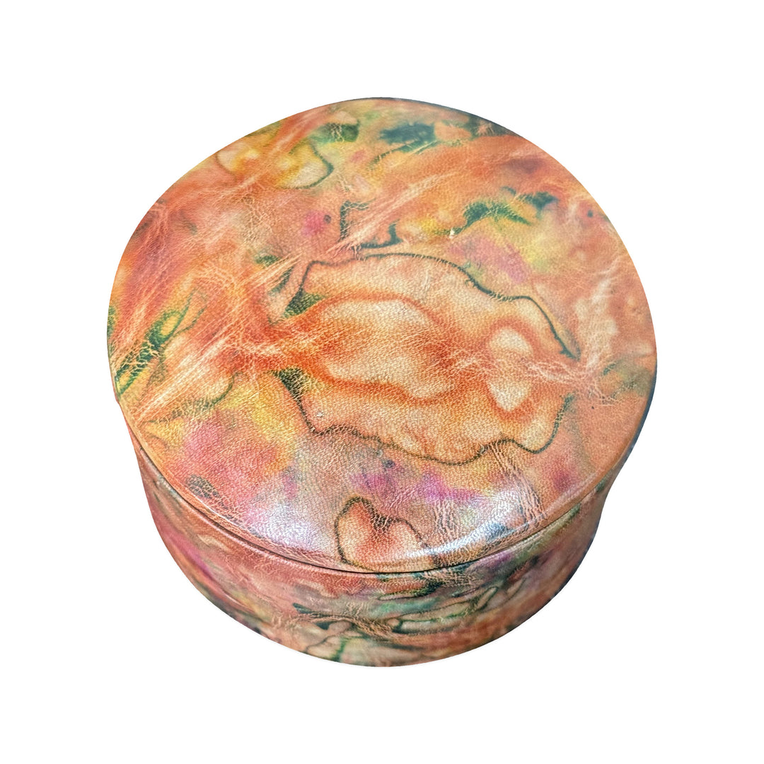 Boîte ronde en cuir marbré corail, orange et vert, fermée et vue de trois quarts sur fond blanc.