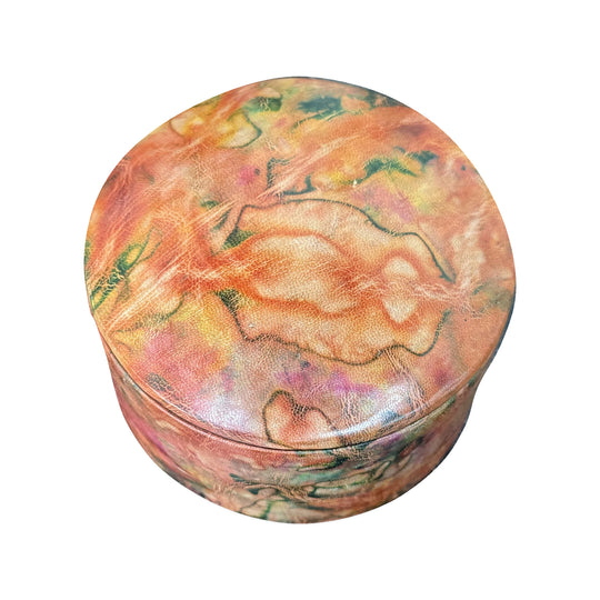 Boîte ronde en cuir marbré corail, orange et vert, fermée et vue de trois quarts sur fond blanc.