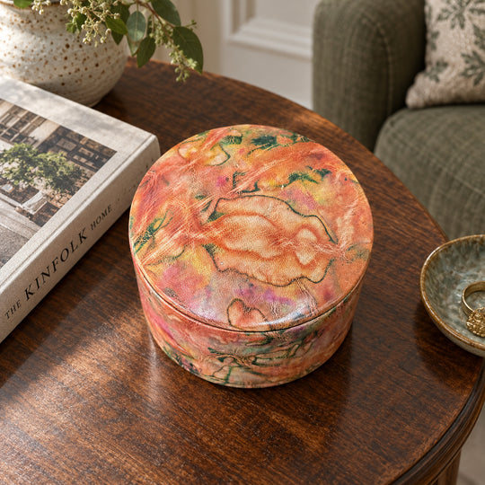 Boîte ronde en cuir marbré corail, orange et vert, sur une table ronde en bois près d'un livre et d'un fauteuil vert.