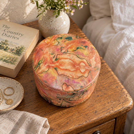 Boîte ronde en cuir marbré corail, orange et vert, sur une table de chevet en bois près d'un livre et d'un lit au linge blanc.