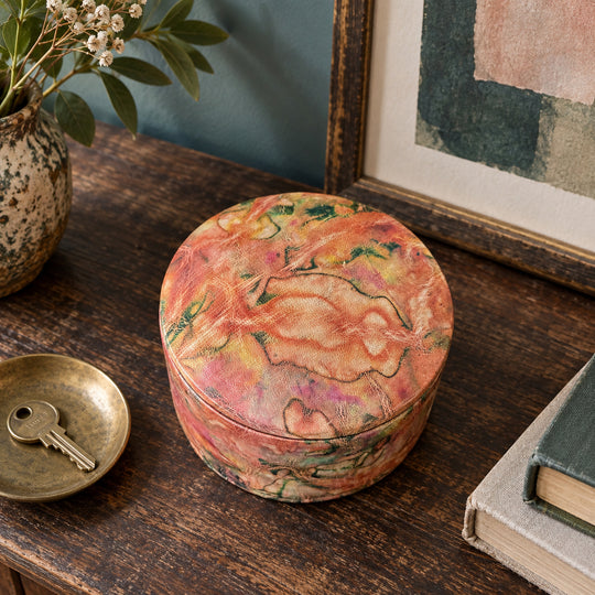Boîte ronde en cuir marbré corail, orange et vert, sur une console en bois devant une œuvre encadrée, près d'une coupelle avec une clé.