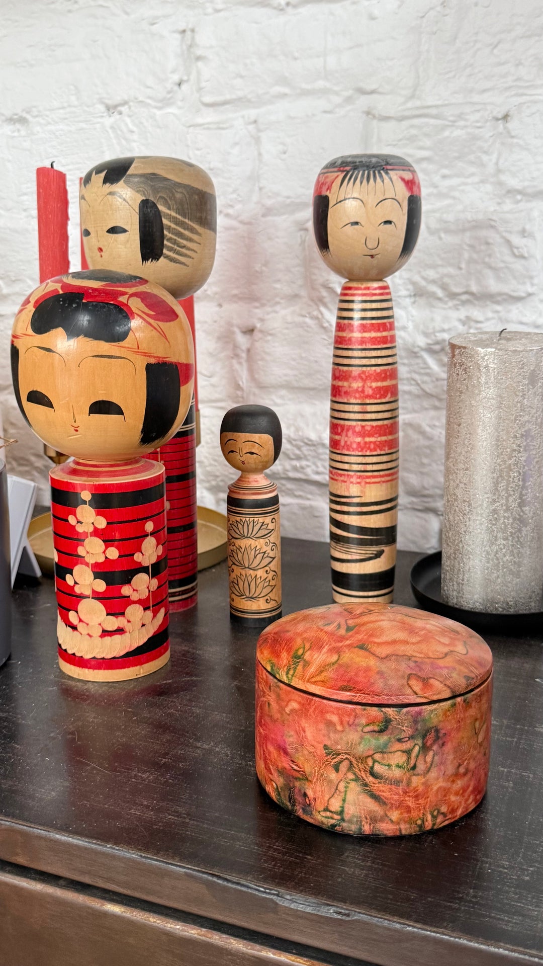Boîte ronde en cuir marbré corail, orange et vert, sur un meuble en bois devant des poupées japonaises kokeshi.