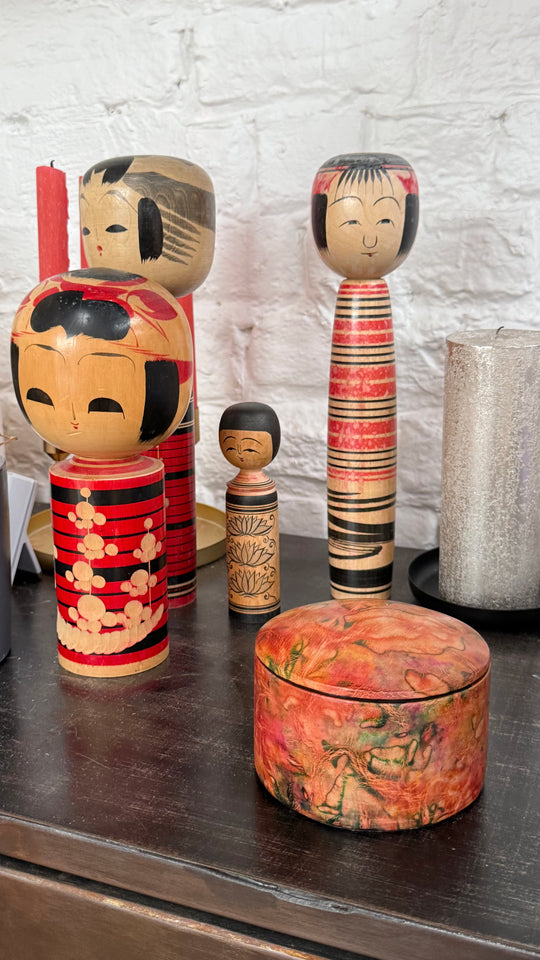 Boîte ronde en cuir marbré corail, orange et vert, sur un meuble en bois devant des poupées japonaises kokeshi.