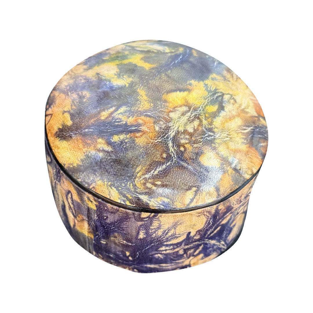 Boîte ronde en cuir marbré violet et jaune ocre, fermée et vue de trois quarts sur fond blanc.