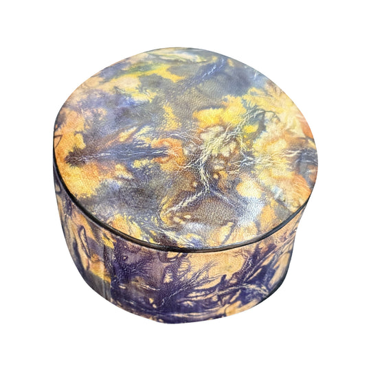 Boîte ronde en cuir marbré violet et jaune ocre, fermée et vue de trois quarts sur fond blanc.