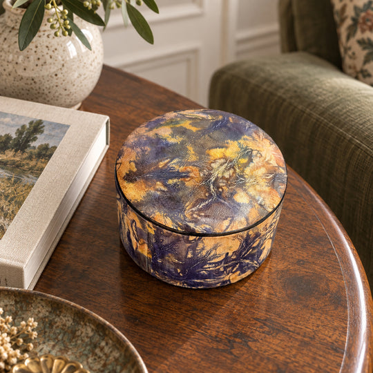 Boîte ronde en cuir marbré violet et jaune ocre, sur une table ronde en bois près d'un livre et d'un fauteuil vert.