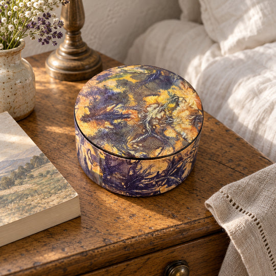Boîte ronde en cuir marbré violet et jaune ocre, sur une table de chevet en bois près d'un livre et d'un lit au linge blanc.