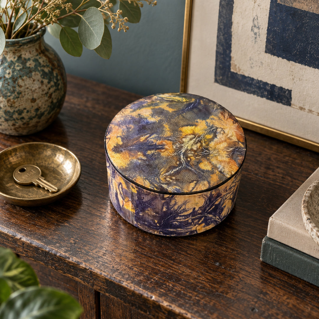 Boîte ronde en cuir marbré violet et jaune ocre, sur une console en bois devant une œuvre encadrée, près d'une coupelle avec une clé.