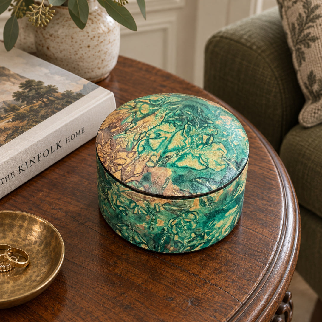 Boîte ronde en cuir marbré vert turquoise et beige, sur une table ronde en bois près d'un livre et d'un fauteuil vert.
