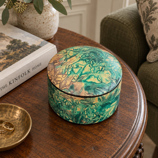 Boîte ronde en cuir marbré vert turquoise et beige, sur une table ronde en bois près d'un livre et d'un fauteuil vert.