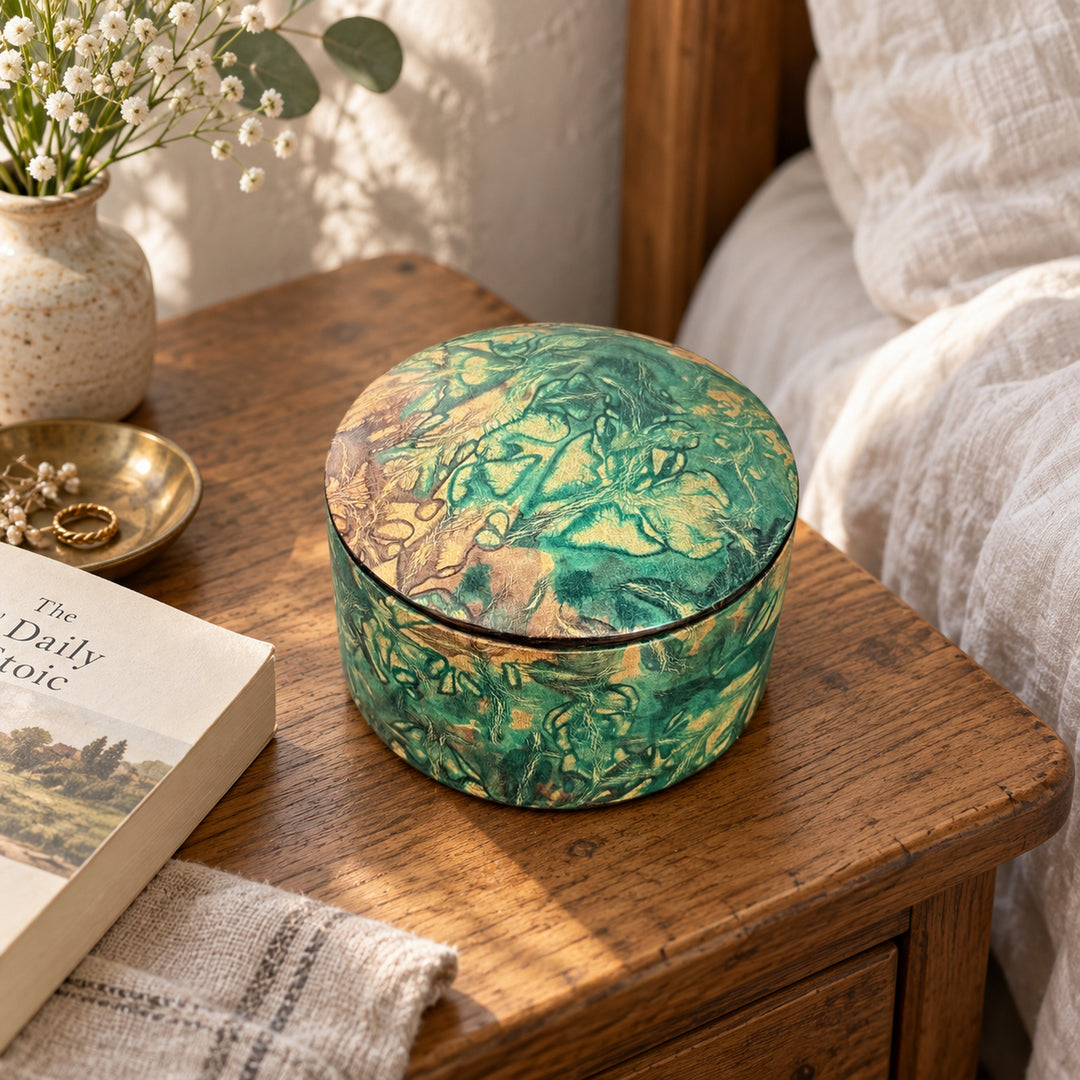 Boîte ronde en cuir marbré vert turquoise et beige, sur une table de chevet en bois près d'un livre et d'un lit au linge blanc.
